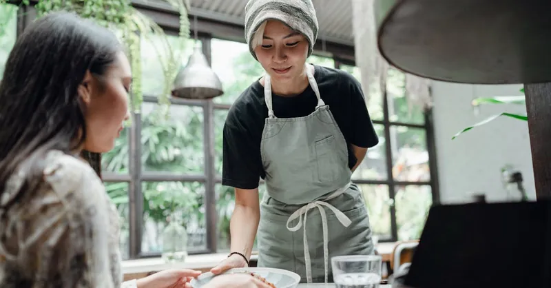 Waitress in uniform serving a meal to a customer in a charming cafe with lush greenery.