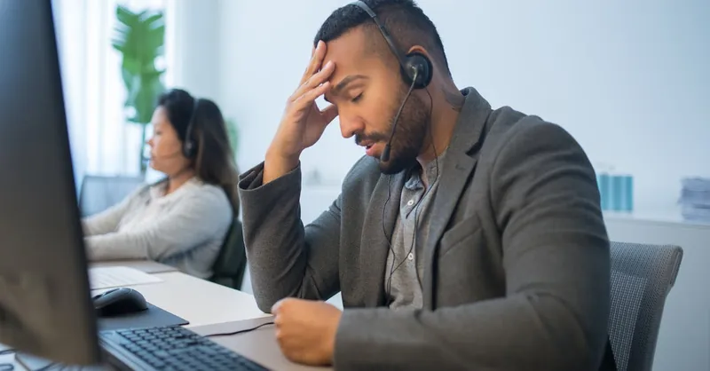 A tired call center employee with a headset looks stressed at his desk in a modern office.