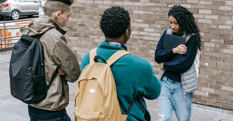 Young multiracial male friends with backpacks offending female with folder on city street near brick wall in daylight