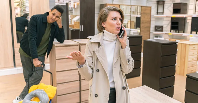 Woman on phone in a furniture store, expressing urgency with a shopping customer nearby.