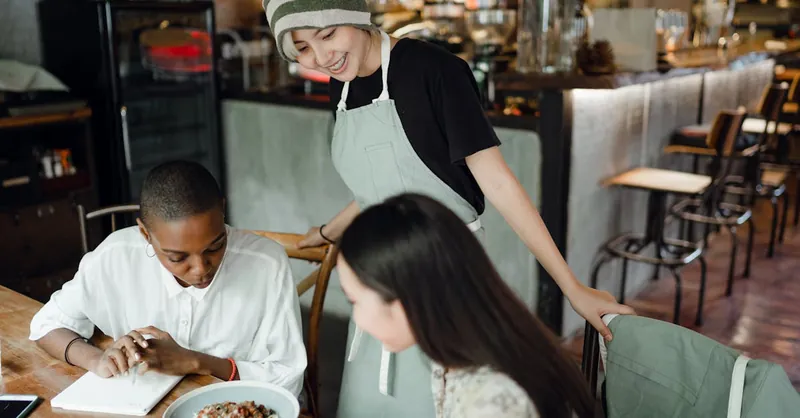 Positive young Asian waitress in apron and hat serving delicious pasta for interested diverse female customers in cozy cafe