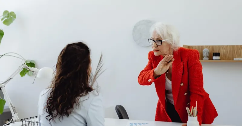 An elderly woman in a red jacket expressing displeasure in an office setting with a colleague.