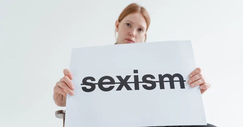 A caucasian woman confidently holds a sign addressing sexism in a bright studio setting.