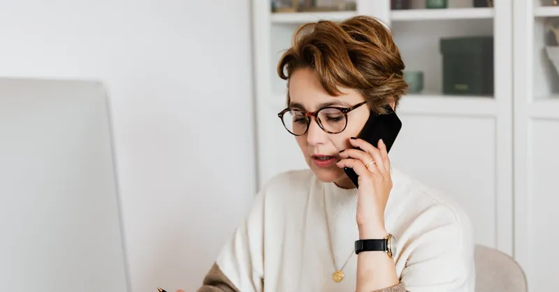 Concentrated short haired female entrepreneur in eyeglasses having discussion via smartphone in modern office