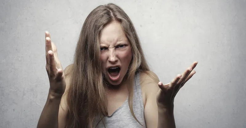A young woman showing intense anger and frustration, yelling with hands raised against a gray background.