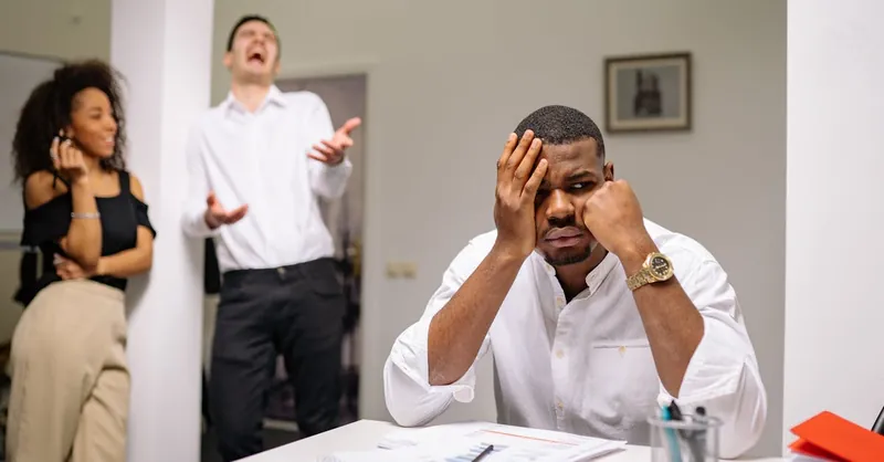 A man at a desk looks frustrated while two coworkers laugh. Office workplace conflict concept.