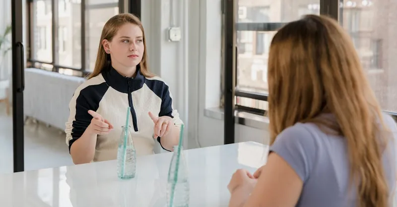 Two women sitting at a table indoors, communicating using sign language, showcasing friendship and inclusivity.