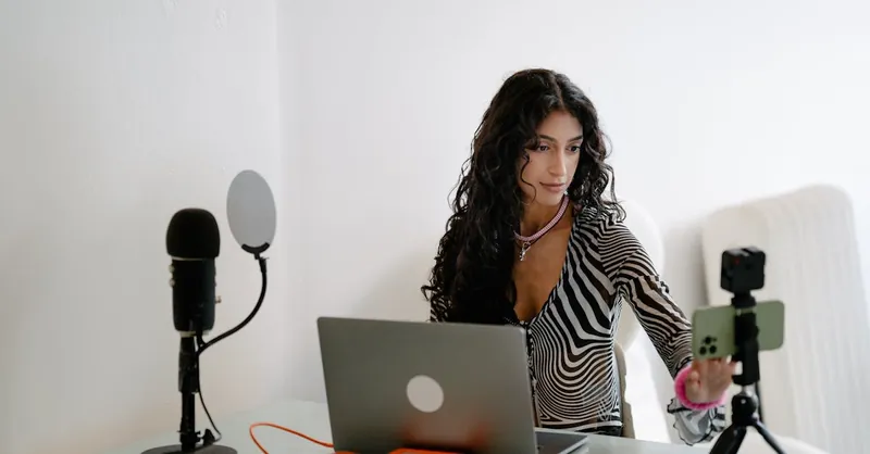 A young woman using a laptop and smartphone for livestreaming, surrounded by tech gadgets indoors.