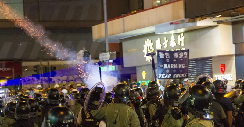 Riot police in tactical gear facing a public protest at night with tear gas.