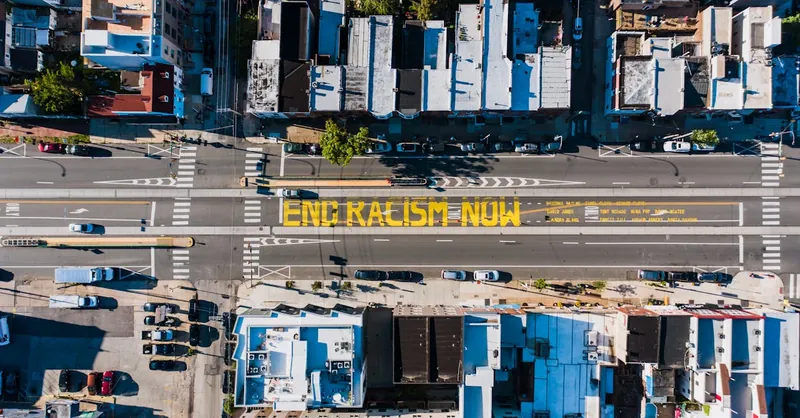 Overhead view of asphalt roadway with vehicles between building roofs and colorful title about racism