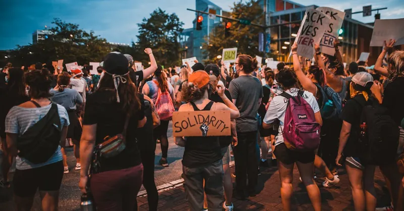 A diverse group of people gathering outdoors with signs advocating for social justice and solidarity.