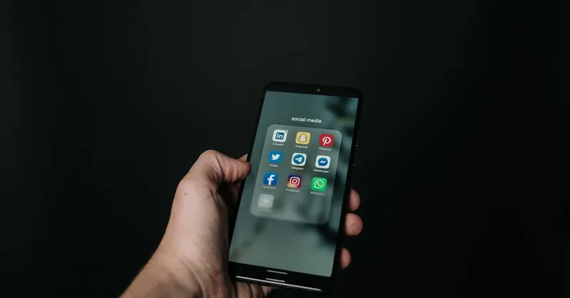 Close-up of a hand holding a smartphone displaying various social media app icons on a dark background.