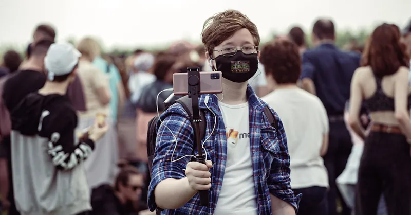 Young individual wearing a mask records a crowd with a smartphone mounted on a selfie stick.